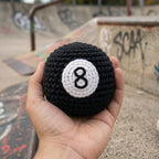 Hand holding a black and white crochet pool ball with number 8 against a graffiti-covered wall.