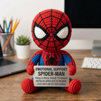 A crocheted Spider-Man doll sits on a wooden desk holding a sign that reads 'EMOTIONAL SUPPORT SPIDER-MAN' with a supportive message. The desk background includes a computer monitor, keyboard, pencil cup, and small plant.
