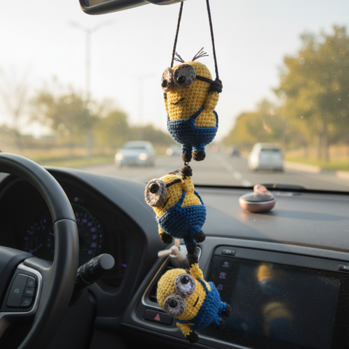 Three crochet minions hanging from a car's rearview mirror with a road and trees in the background.